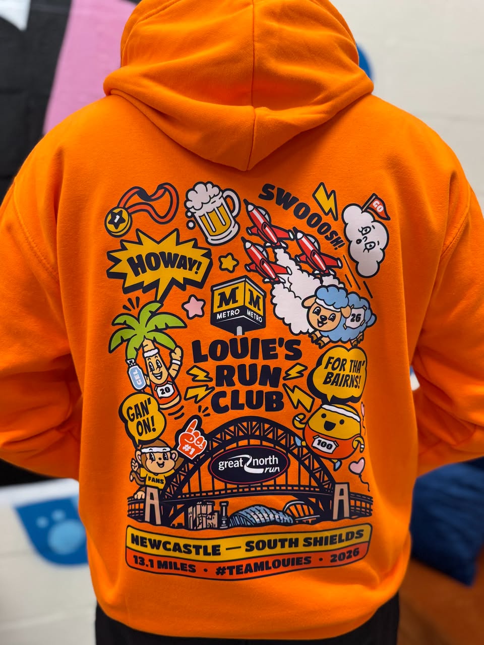 A person viewed from behind wearing a bright orange hoodie with a large colorful back graphic. The graphic includes cartoon characters, a beer mug, a rocket, the text 'LOUIE'S RUN CLUB,' 'NEWCASTLE — SOUTH SHIELDS,' '12.